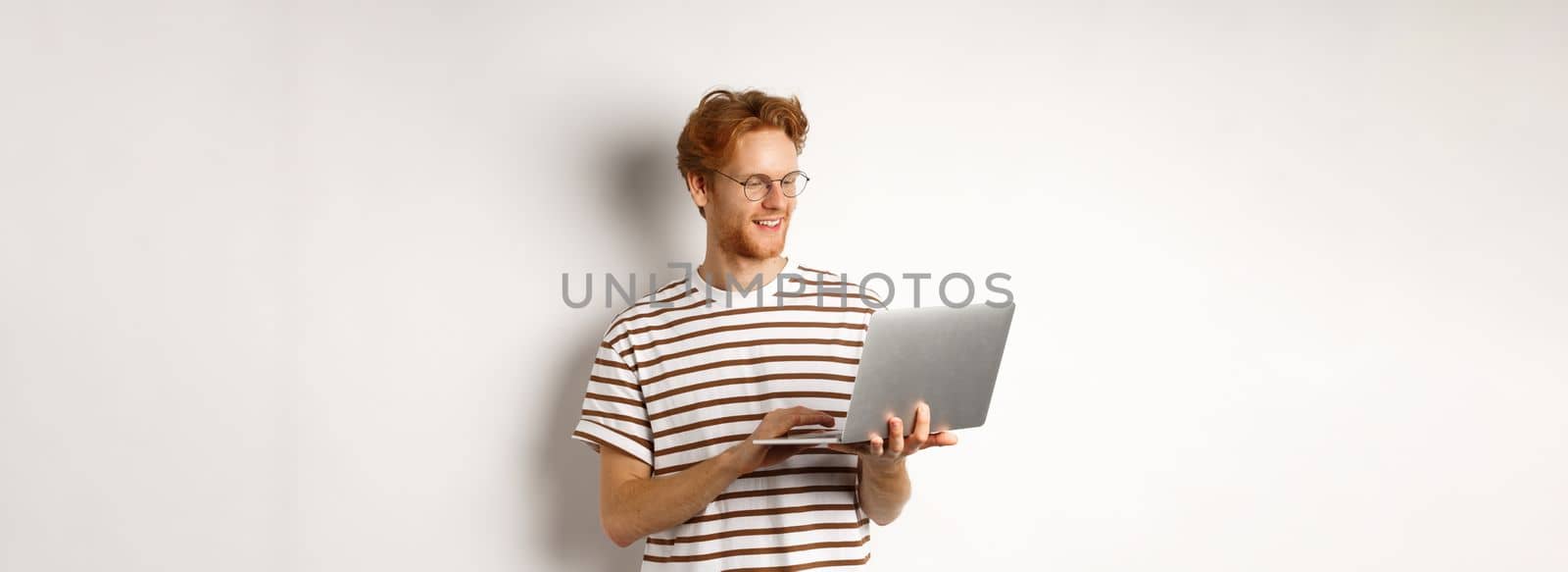 Young redhead freelancer working on laptop, typing on computer keyboard and smiling, standing over white background.