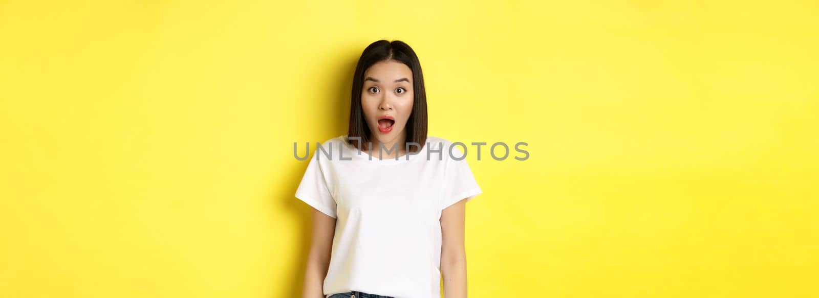 Beauty and fashion concept. Surprised asian girl open mouth and staring at camera, drop jaw impressed, standing over yellow background.
