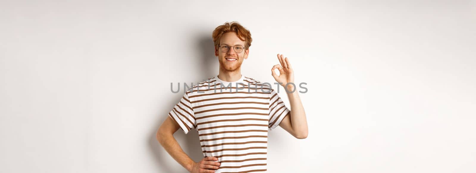 Confident smiling man with red hair assuring you, showing OK sign, guarantee quality, recommending something good, standing over white background.