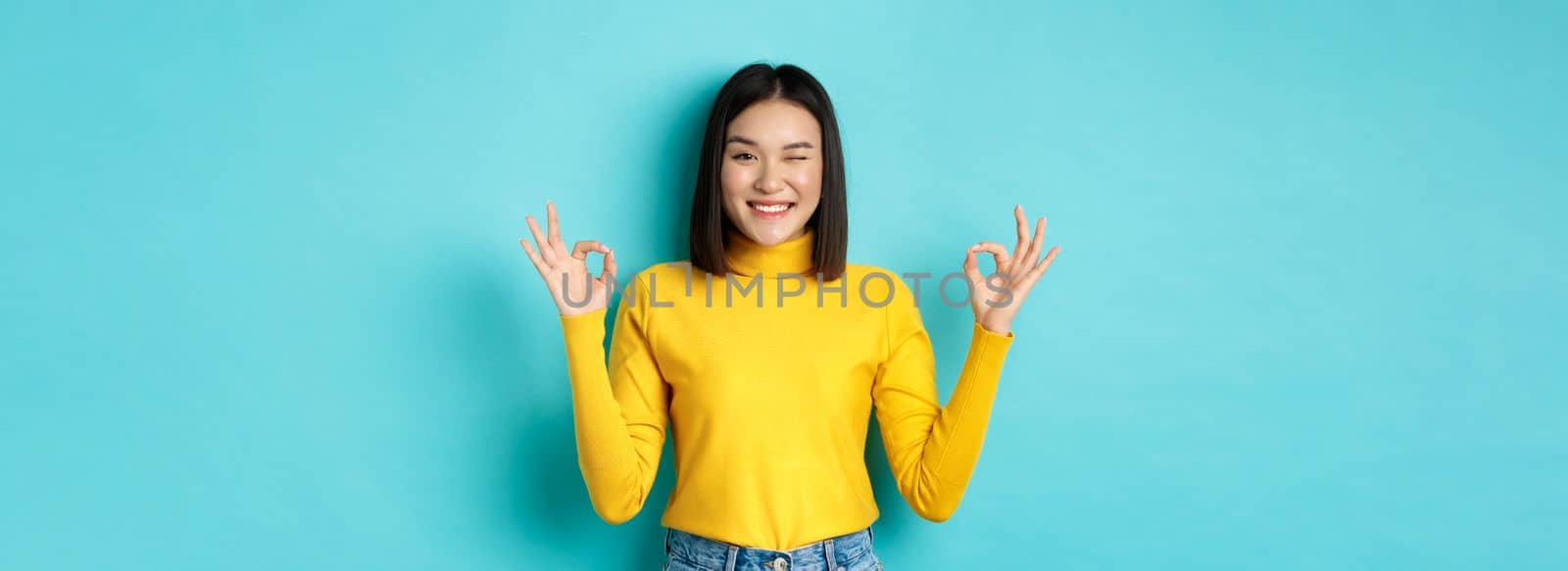 Cheerful asian female model showing okay gestures, smiling and looking impressed, praise product, standing over blue background.