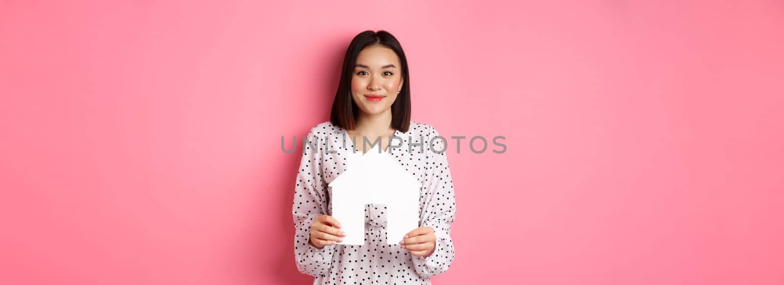Real estate. Adult asian woman searching for home, holding house model and smiling, promo of broker company, standing over pink background by Benzoix