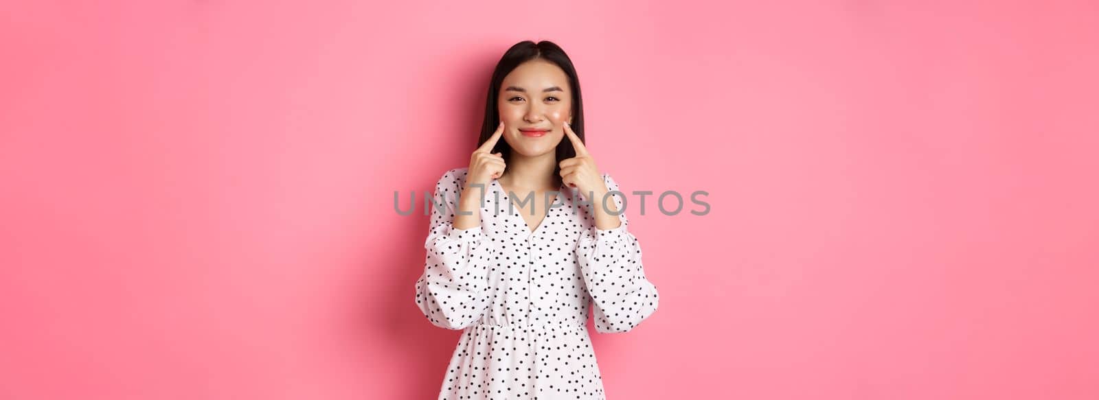 Cute blushing asian girl poking cheeks, smiling happy at camera, wearing romantic white dress, standing over pink background.