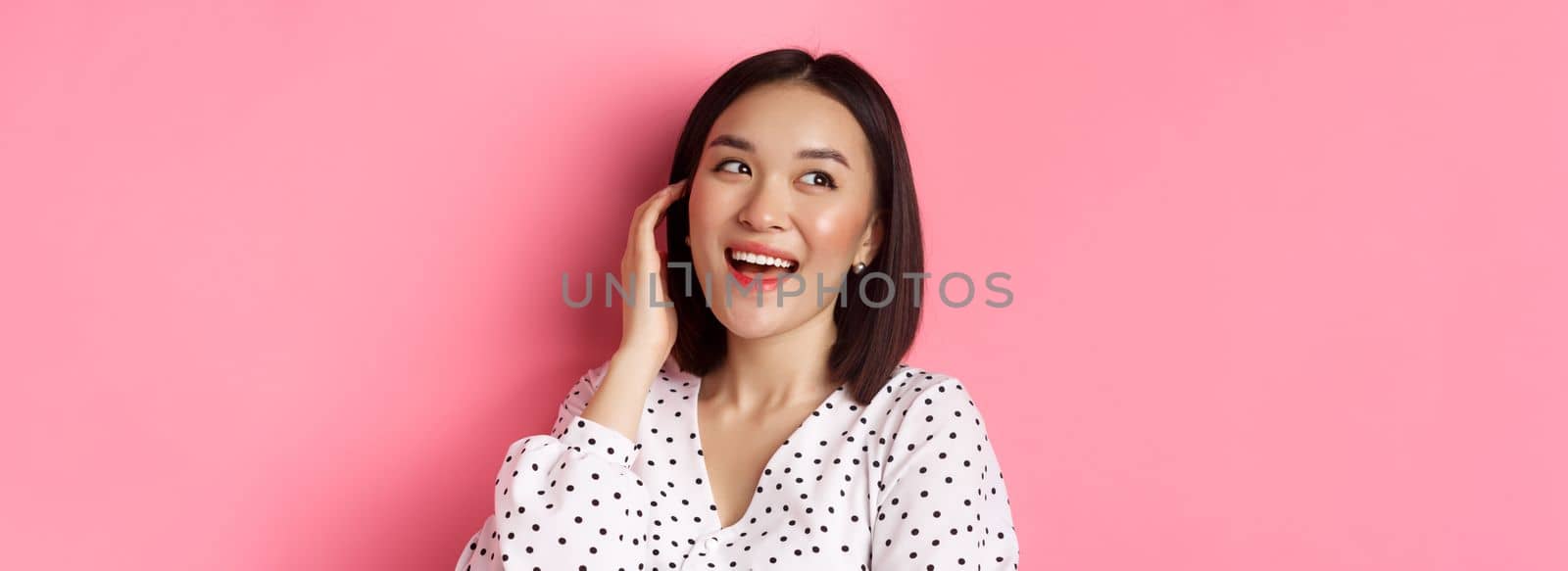 Close-up of flirty asian woman sighing, looking dreamy left and smiling, standing over pink background by Benzoix