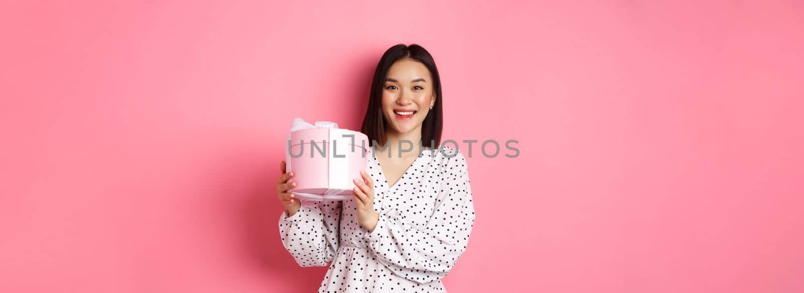 Romantic asian woman in cute dress holding box with gift, smiling happy at camera, standing with present over pink background.