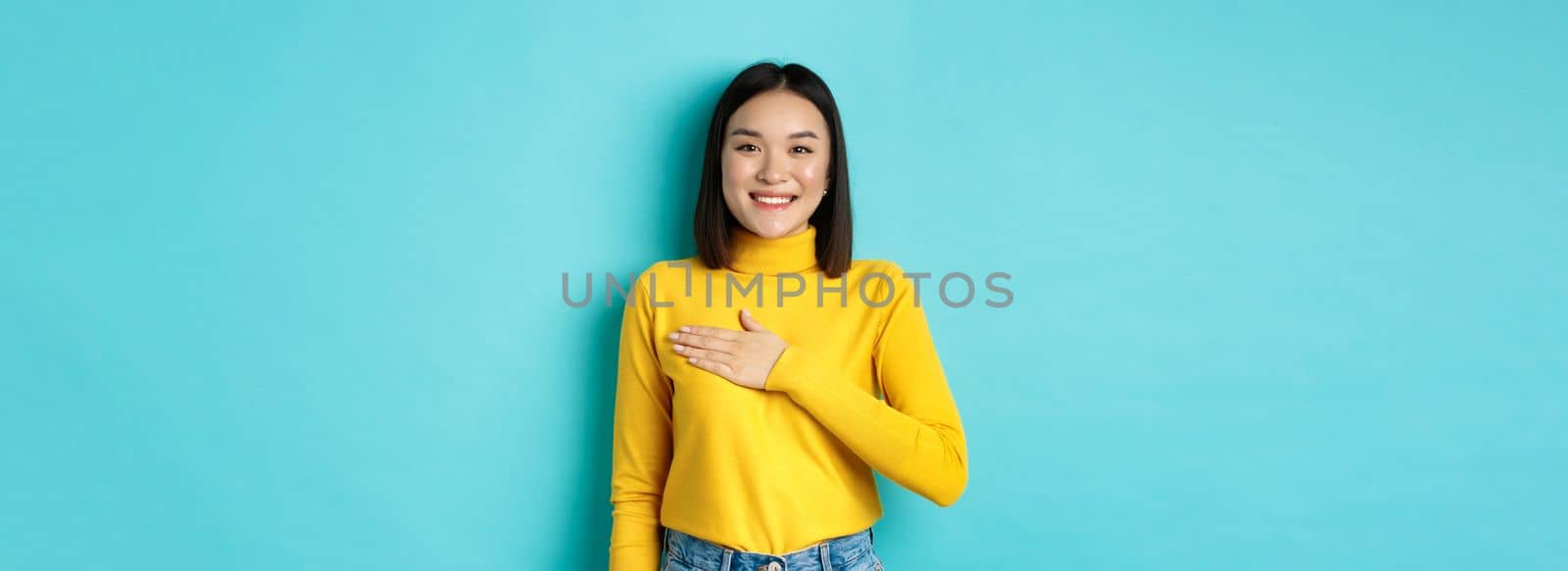 Image of proud smiling asian woman holding hand on heart, showing respect to national anthem, standing over blue background. Copy space