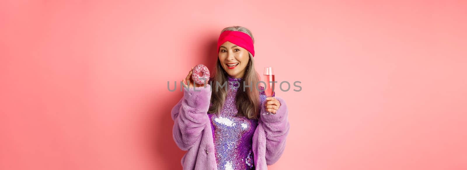 Beautiful and stylish asian senior woman having fun, drinking champagne and eating sweet donut, smiling at camera, standing over pink background.