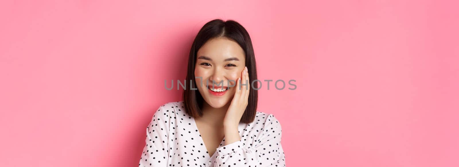 Close-up of adorable asian woman blushing, touching cheek and smiling cute at camera, standing over pink background by Benzoix