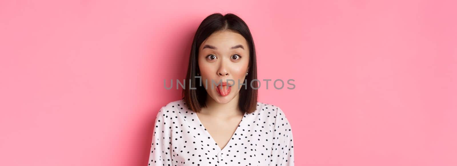 Beauty and lifestyle concept. Close-up of funny and cute asian woman showing tongue, staring at camera silly, standing over pink background by Benzoix