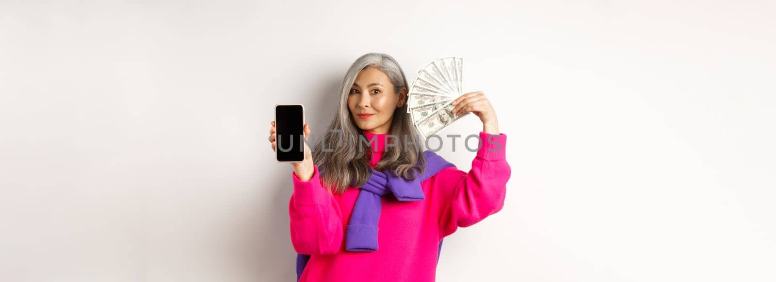 Fashionable asian senior woman showing money dollars and blank smartphone screen, demonstrate online shop, standing over white background.