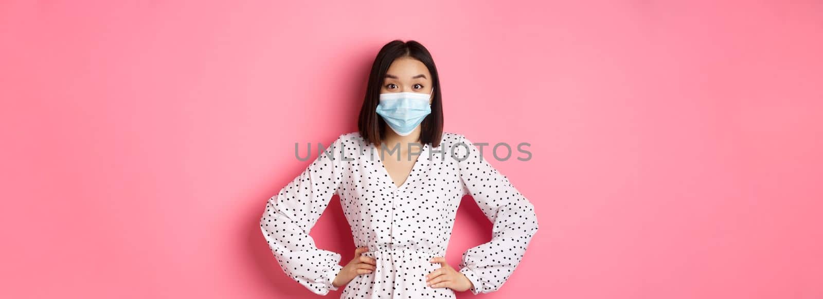 Covid-19, quarantine and lifestyle concept. Cute korean woman in dress and face mask using preventive measures from coronavirus, standing over pink background.