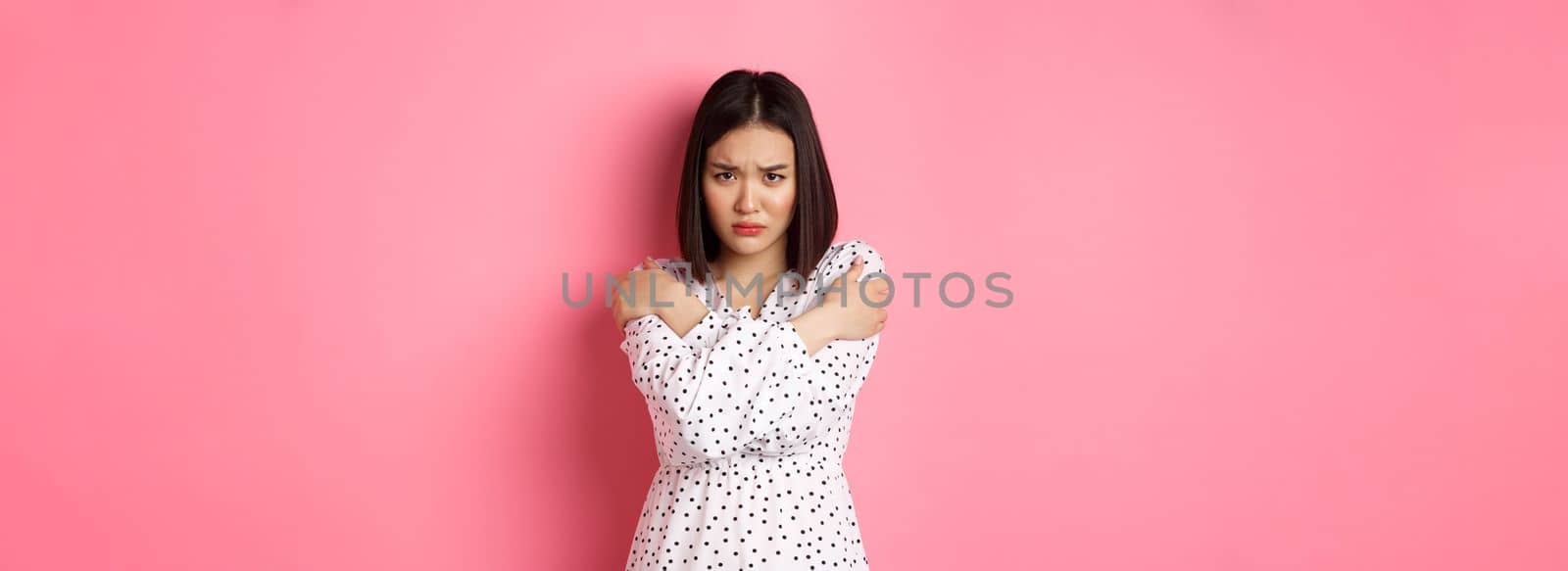 Timid and offended asian girl cross arms on chest, staring defensive and insulted at camera, standing in dress over pink background by Benzoix