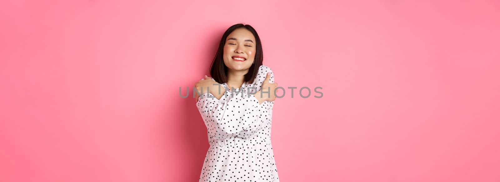 Beautiful asian woman feeling romantic, hugging herself, close eyes and smiling with dreamy face, standing happy over pink background by Benzoix