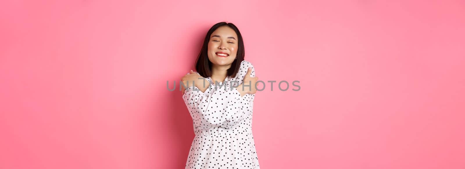 Beautiful asian woman feeling romantic, hugging herself, close eyes and smiling with dreamy face, standing happy over pink background.