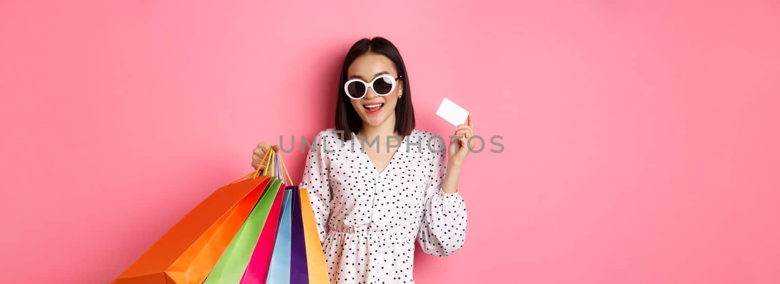 Beautiful asian woman in sunglasses going shopping, holding bags and showing credit card, standing over pink background by Benzoix