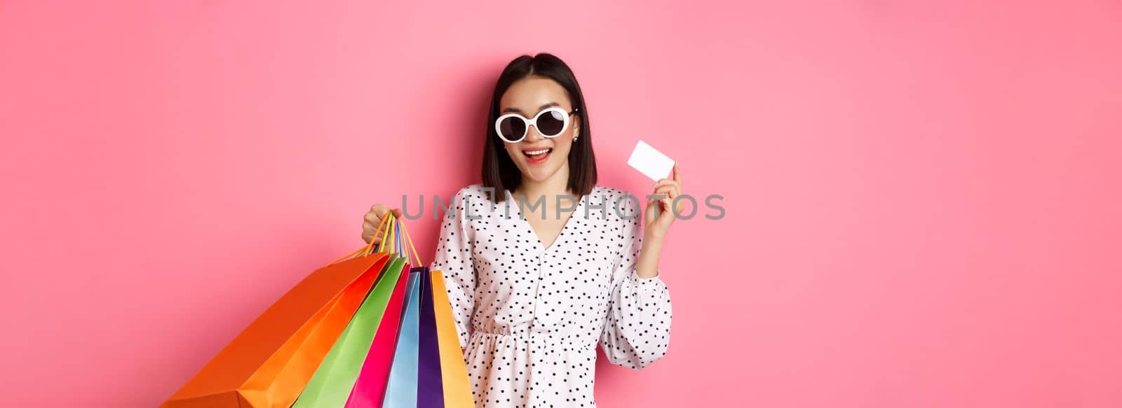 Beautiful asian woman in sunglasses going shopping, holding bags and showing credit card, standing over pink background.