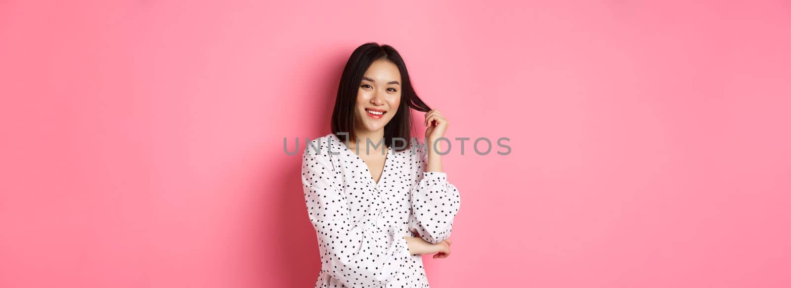 Cute asian female brunette in trendy dress, playing with hair and smiling flirty, standing in dress over pink background.