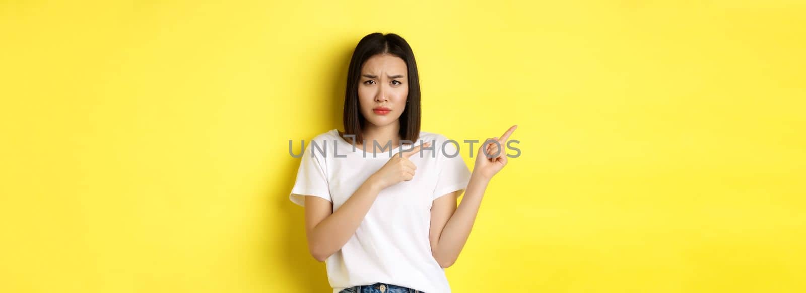 Beauty and fashion concept. Beautiful asian woman in white t-shirt pointing fingers left, standing over yellow background.