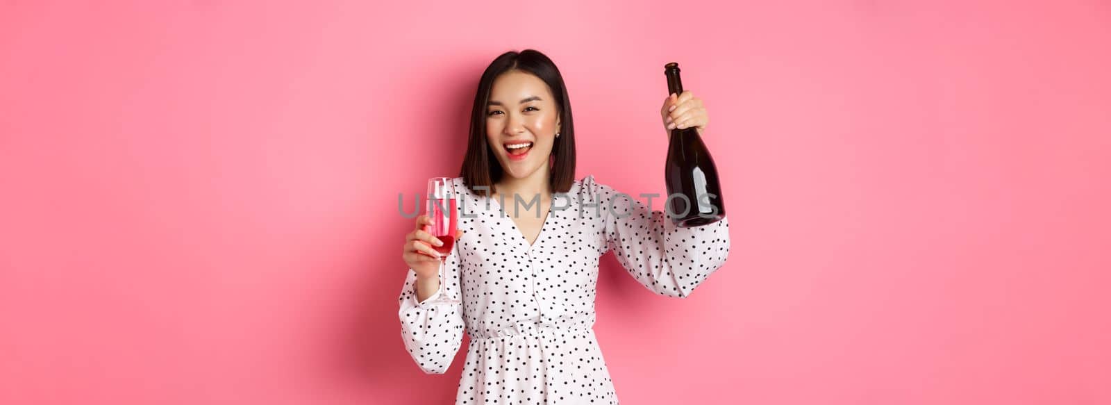 Happy asian woman celebrating, having fun and partying, pouring glass of champagne and laughing, standing over pink background by Benzoix