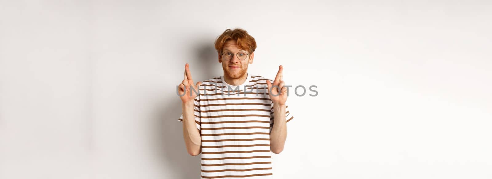 Hopeful young man with red hair and glasses cross fingers for good luck, making wish, standing over white background.