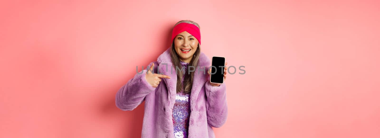 Online shopping and fashion concept. Stylish asian mature lady showing blank smartphone screen, pointing at phone and smiling, pink background.