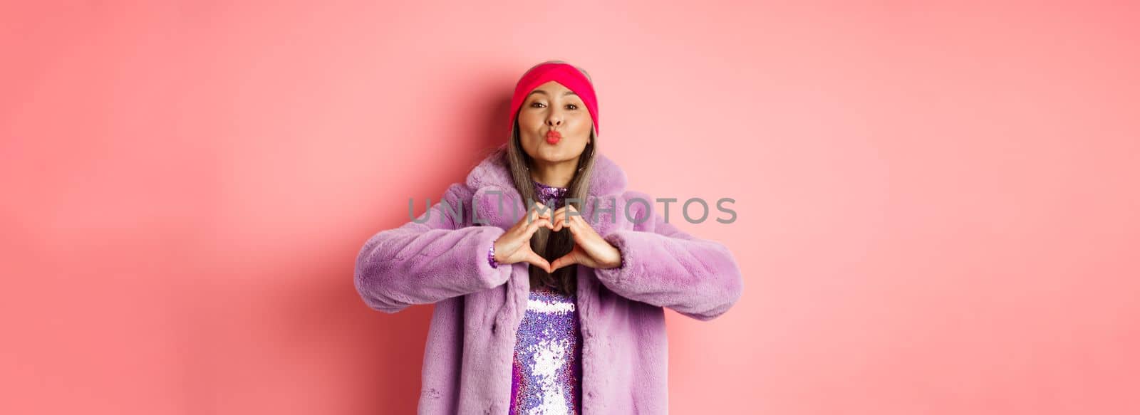 Romance and valentines day. Happy asian senior woman showing heart sign, I love you gesture, pucker lips for kiss, standing over pink background.