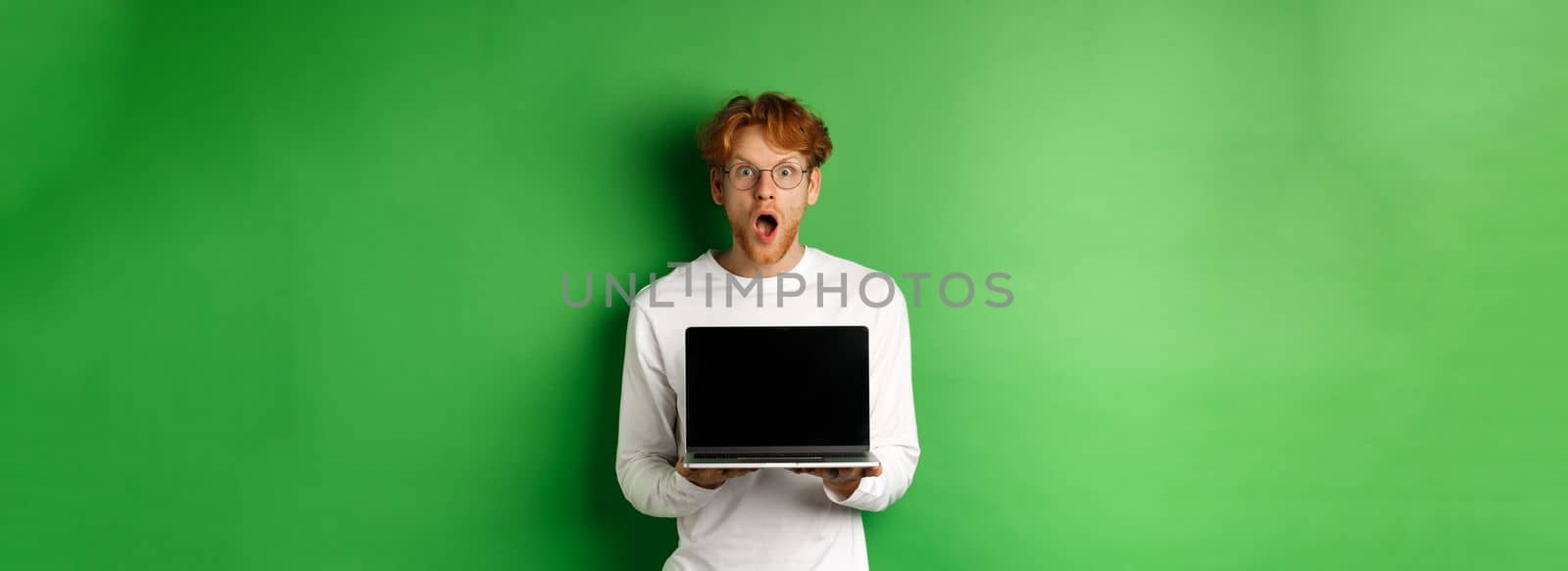 Surprised redhead man in glasses showing laptop blank screen, showing online promo and drop jaw, staring at camera amazed, green background.