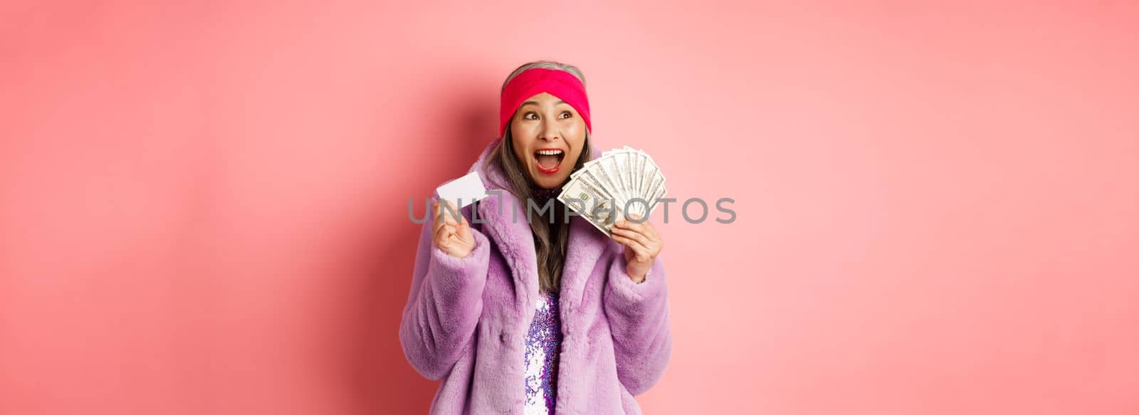 Shopping and fashion concept. Asian senior woman scream happy like winner, holding dollars money and plastic credit card, looking excited, pink background.