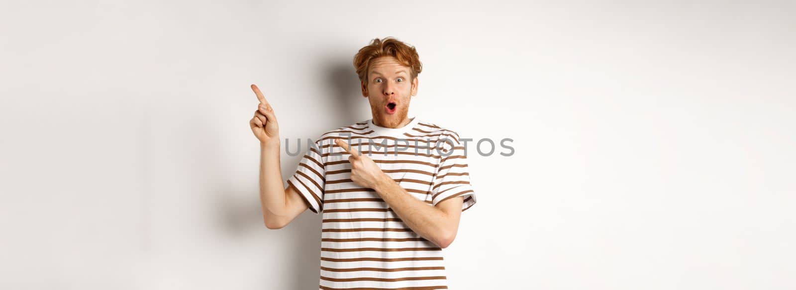 Excited man with curly red hair showing promo offer and looking amazed, pointing fingers at upper right corner logo, white background.