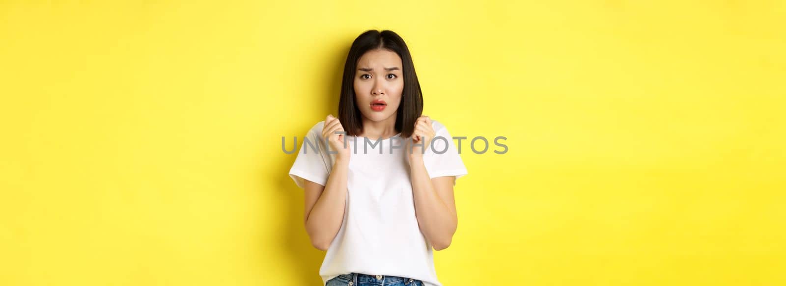 Beauty and fashion concept. Scared timid asian girl looking anxious, gasping startled and standing over yellow background.