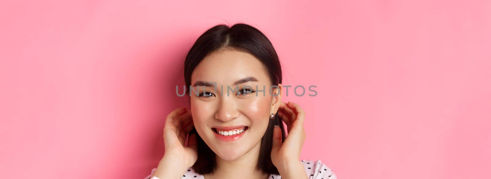 Beauty and skin care concept. Close-up of adorable smiling asian woman tuck hair behind ears, blushing and gazing at camera, standing over pink background.