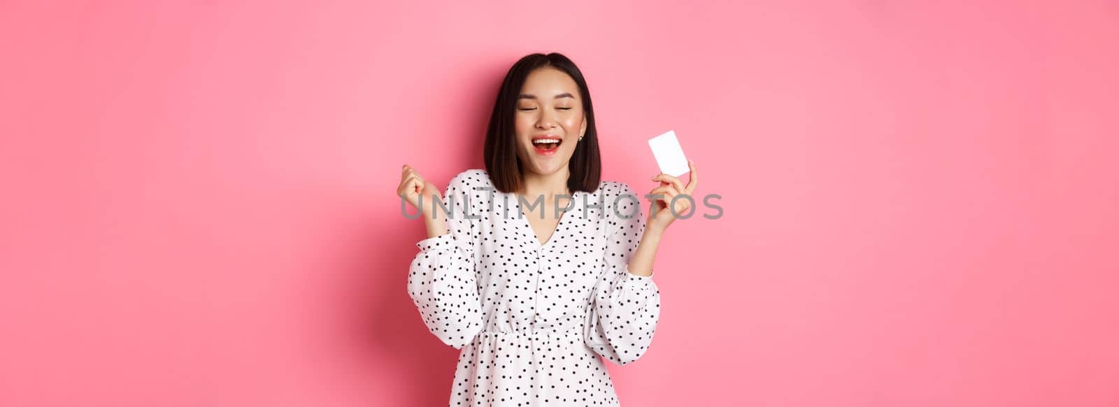 Shopping concept. Excited asian woman feeling satisfaction, holding credit card and rejoicing, standing over pink background.