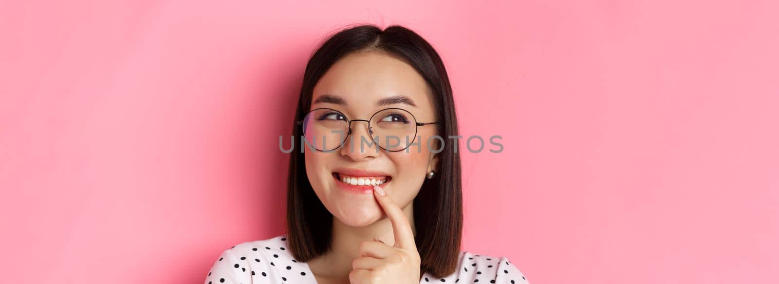 Headshot of cute asian woman in trendy glasses smiling, having an idea, thinking and looking at upper left corner, pink background.