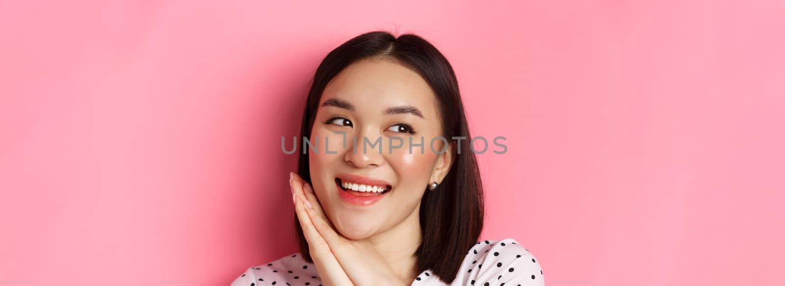 Beauty and skin care concept. Headshot of adorable and dreamy asian woman looking left, smiling and imaging, standing against pink background.
