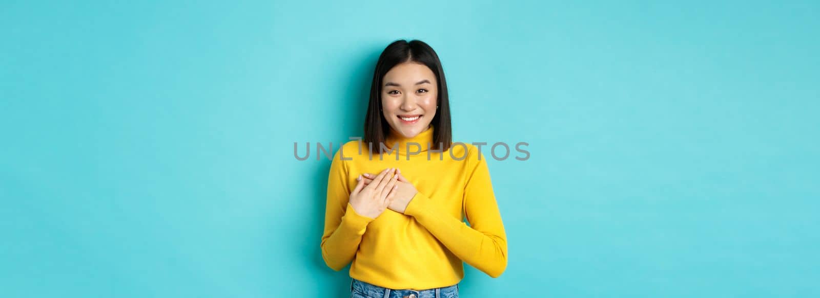 Image of beautiful asian woman holding hands on heart and smiling, thanking you, feeling grateful, standing over blue background by Benzoix