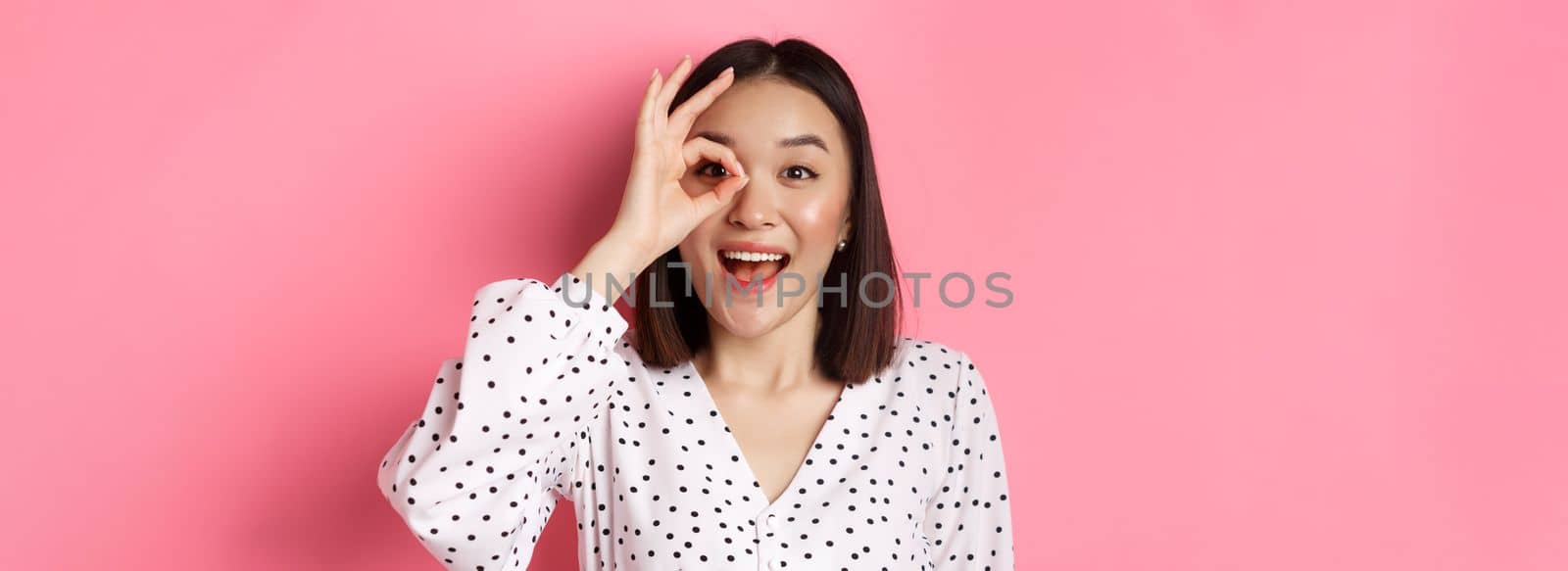 Beauty and lifestyle concept. Beautiful asian girl looking through okay sign and smiling amazed, standing against pink background.
