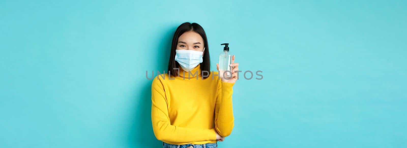 Covid-19, social distancing and pandemic concept. Cheerful asian woman showing hand sanitizer, recommend use antiseptic from coronavirus, wearing medical mask.