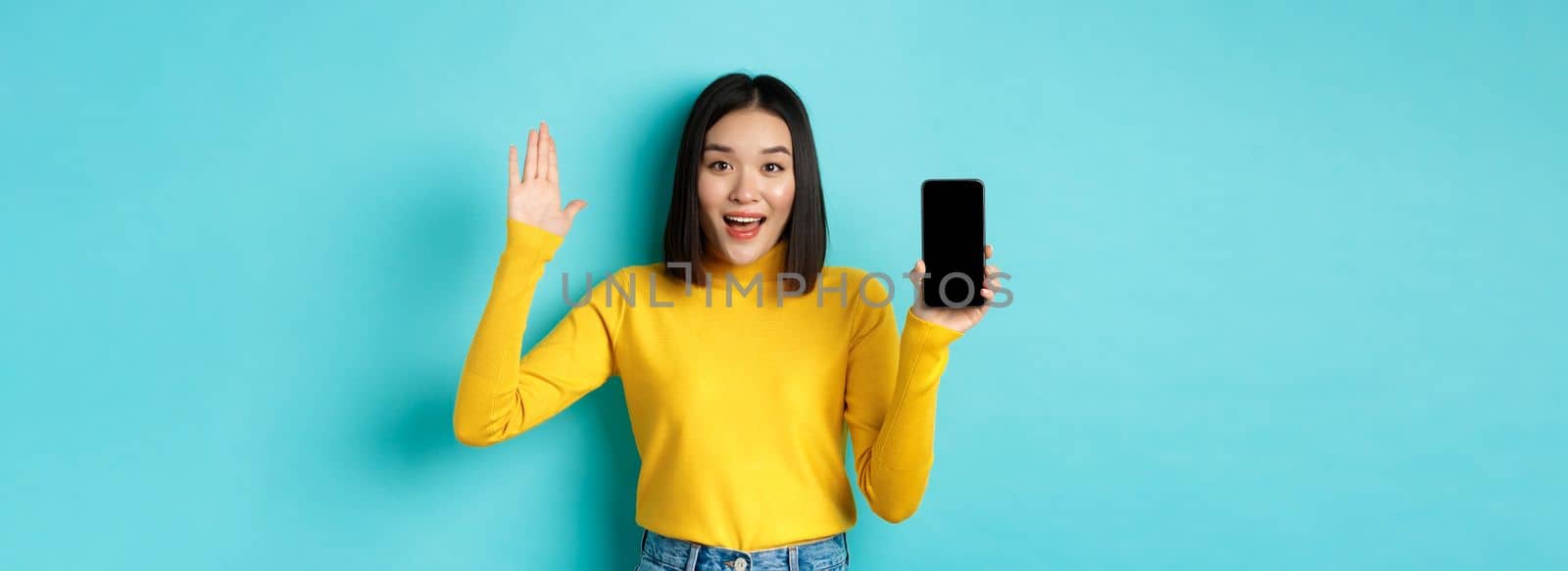 Cheerful asian woman showing empty smartphone screen and raising hand to say hello, greeting you, standing against blue background.