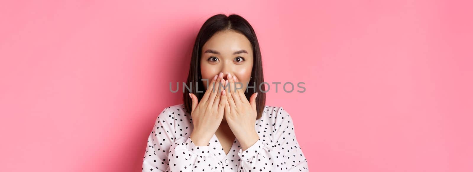 Close-up of beautiful asian girl looking surprised and excited, cover mouth and staring amazed at camera, standing over pink background by Benzoix