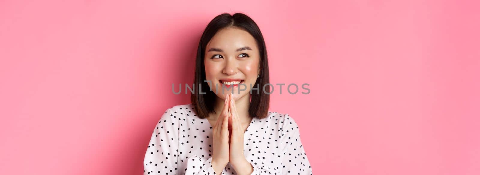Beauty and lifestyle concept. Close-up of happy and satisfied asian woman clap hands, expect something good, smiling and looking aside, standing over pink background.