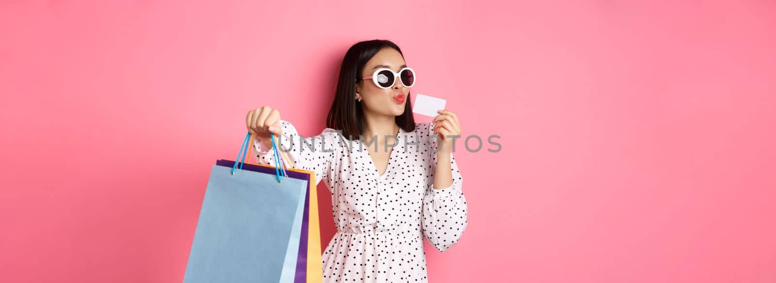 Beautiful asian woman in sunglasses going shopping, holding bags and kissing credit card, standing over pink background by Benzoix