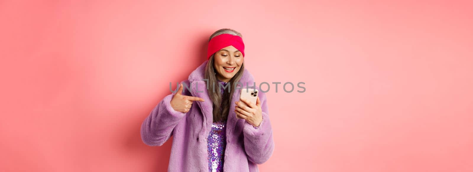 Online shopping and fashion concept. Cool asian senior woman checking out promotion on mobile phone, pointing finger at smartphone screen and smiling pleased, pink background.