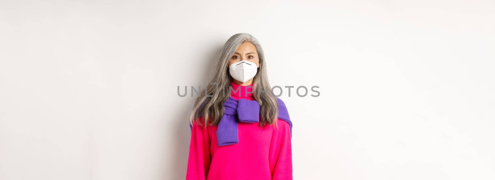 Covid, pandemic and social distancing concept. Stylish asian senior woman wearing respirator and looking at camera serious, standing over white background.