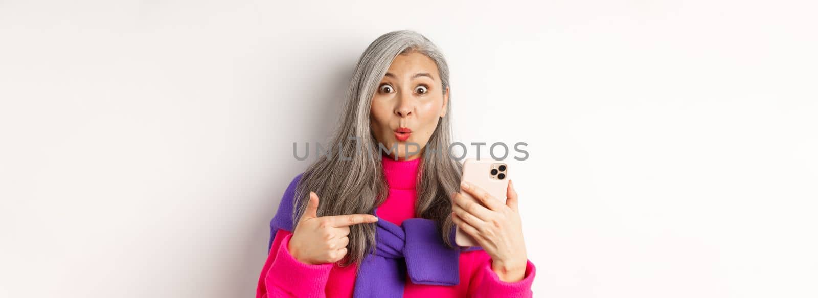 Online shopping. Close up of amazed asian senior woman pointing at smartphone and smiling amazed, standing over white background.