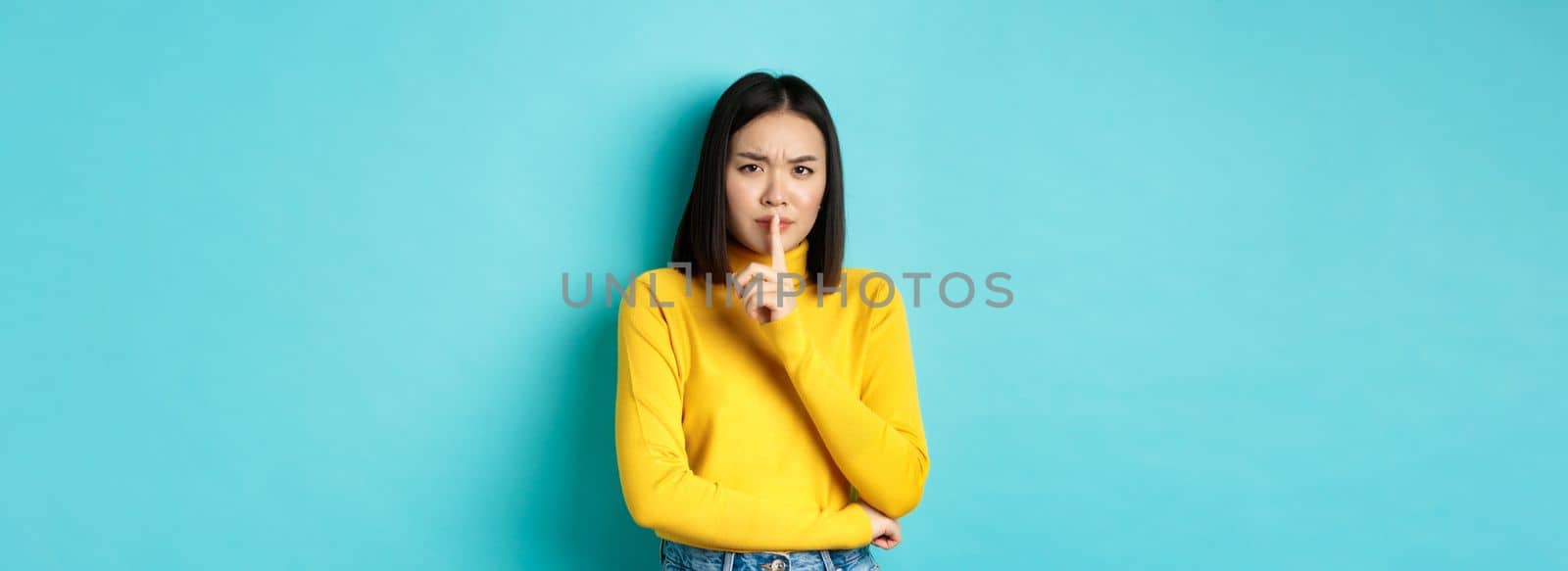 Disappointed asian woman telling to be quiet, scolding loud person with hush gesture, shushing at camera and frowning upset, standing over blue background.