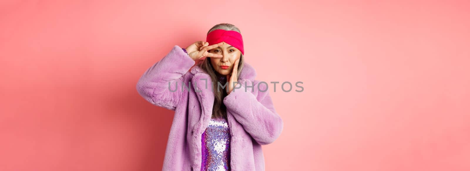 Fashion and shopping. Beautiful asian senior woman in stylish faux fur coat and headband, making peace sign on face and looking sassy and confident at camera, pink background.