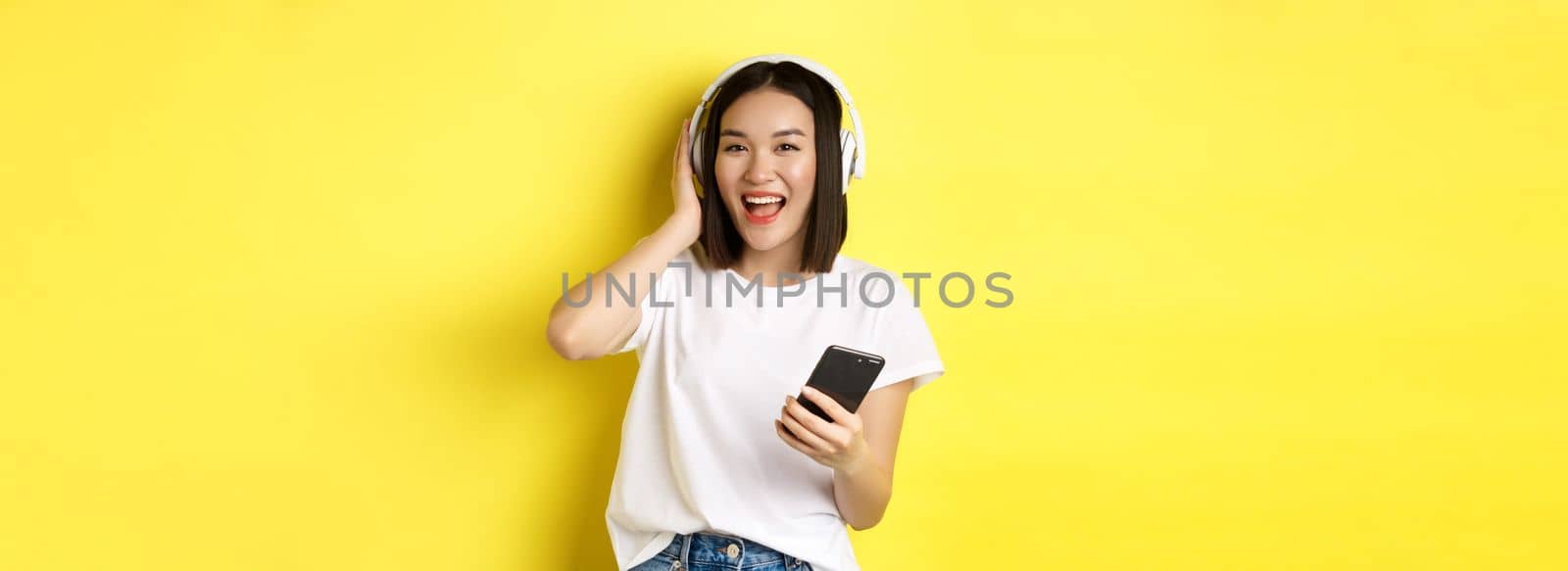 Cool asian girl dancing and listening music in wireless headphones, holding smartphone in hand, standing over yellow background.