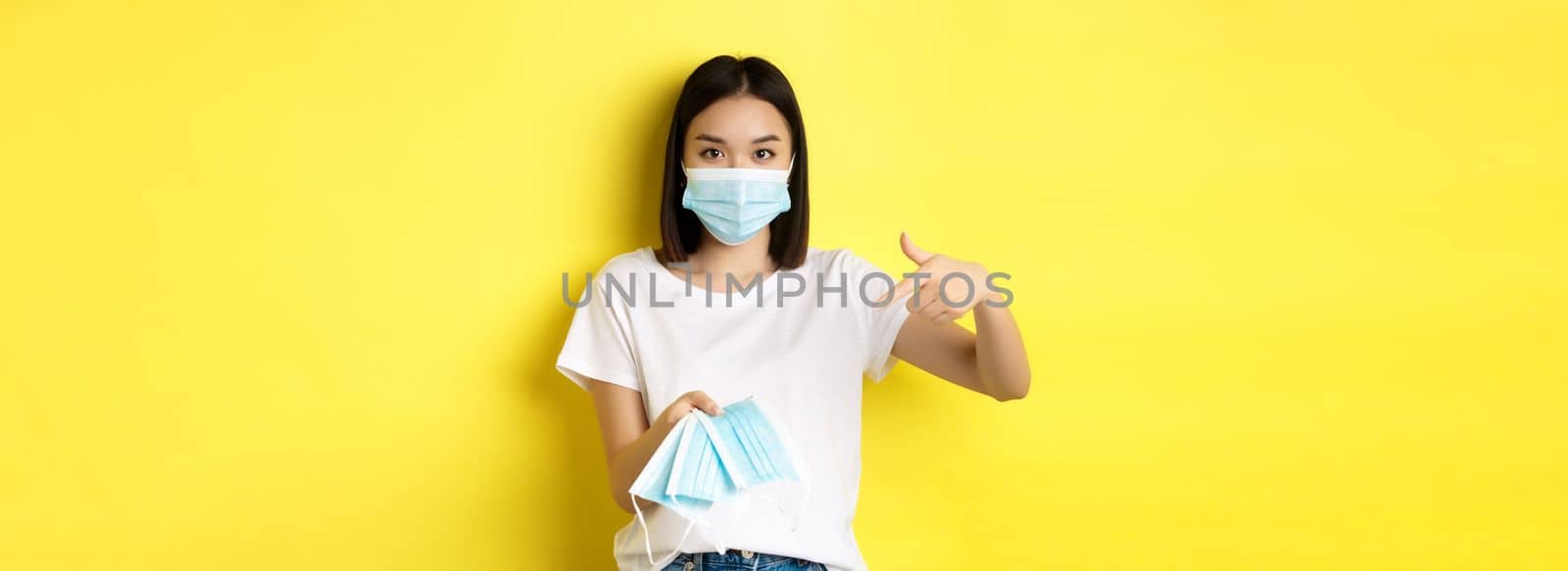 Coronavirus, quarantine and medicine concept. Young asian woman pointing finger at medical masks for going outdoors, standing over yellow background.