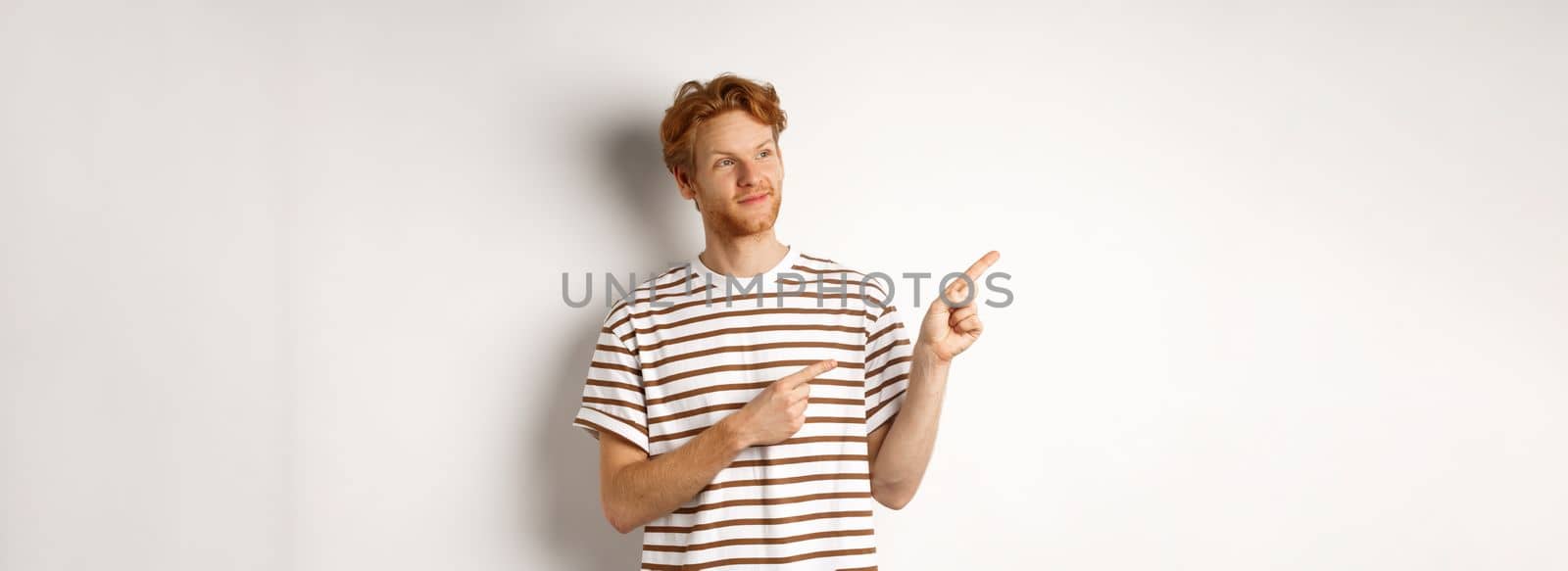 Smiling man with curly red hair, wearing striped t-shirt, smiling and pointing fingers left, demonstrate banner, standing over white background.