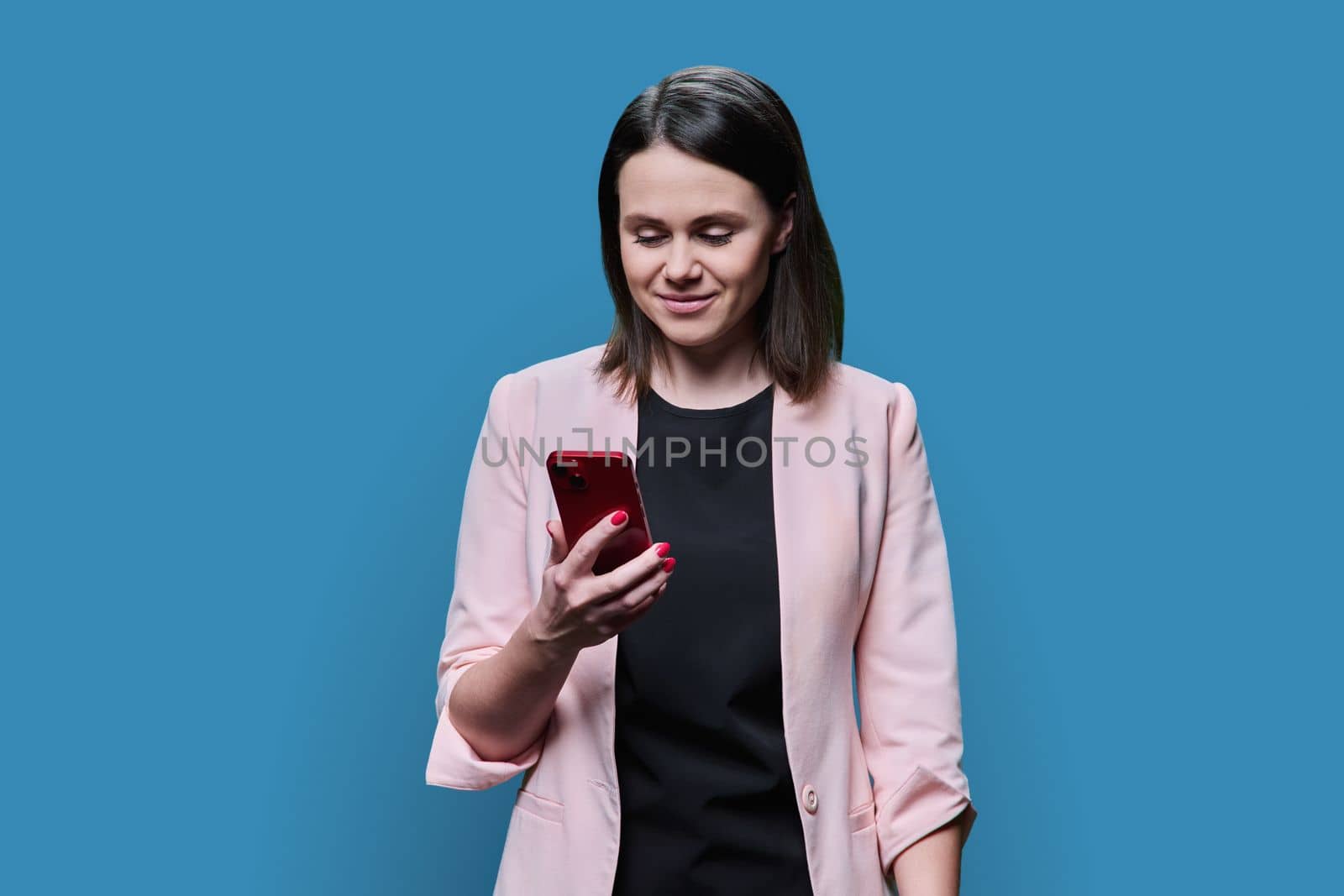 Young business woman using smartphone on blue color background. Positive smiling female in glasses jacket looking texting on phone. Mobile applications, digital technologies in business work leisure