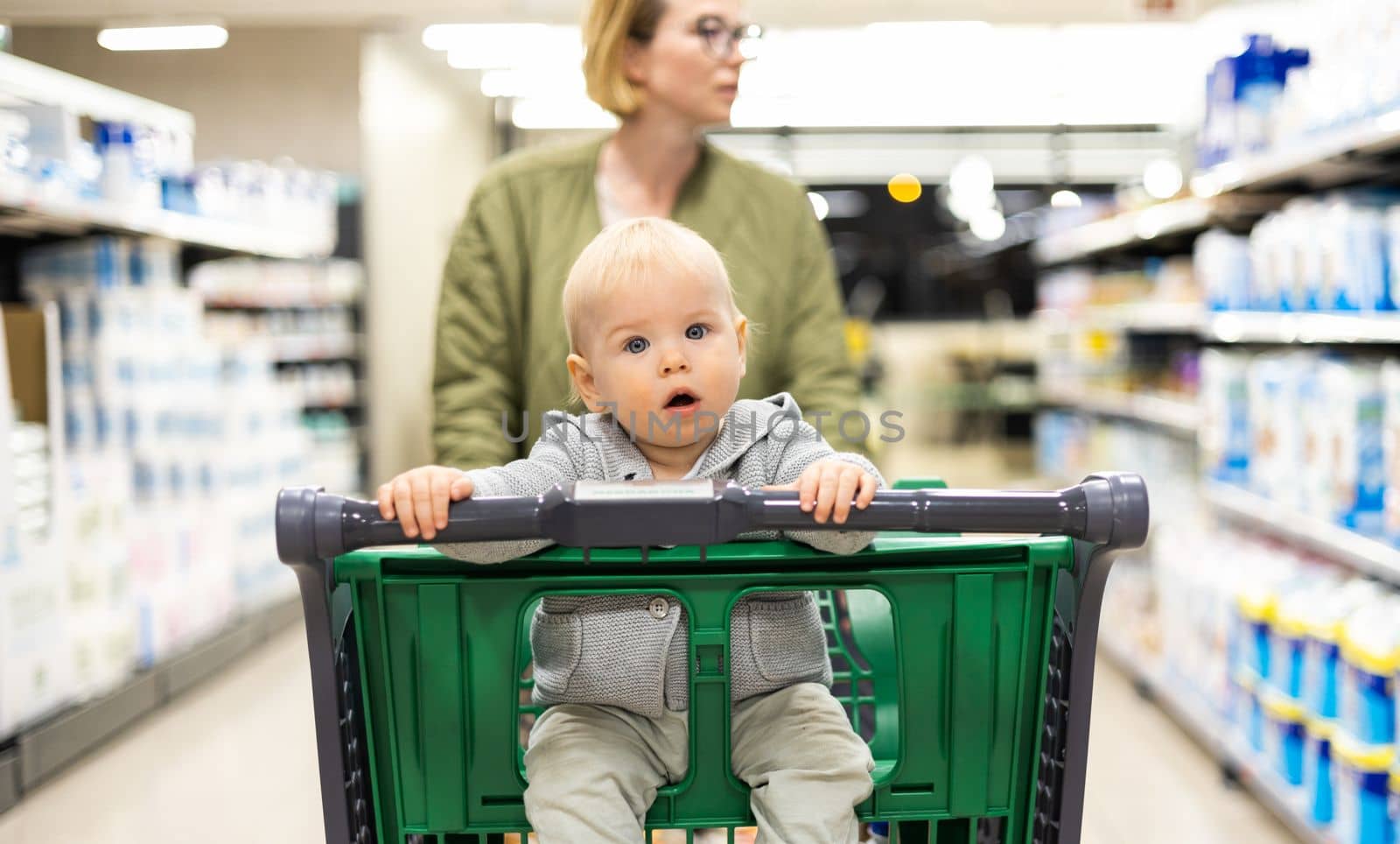 Mother pushing shopping cart with her infant baby boy child down department aisle in supermarket grocery store. Shopping with kids concept